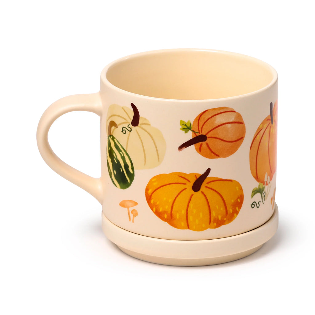Angled view of a cream porcelain mug with its handle visible, featuring an autumn harvest design of pumpkins, gourds, and mushrooms, resting on a matching coaster base.