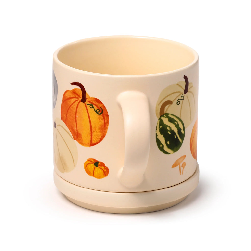 Angled view of a cream porcelain mug with a handle, featuring an autumn harvest pumpkin and gourd design, with its matching coaster lid integrated as the base.