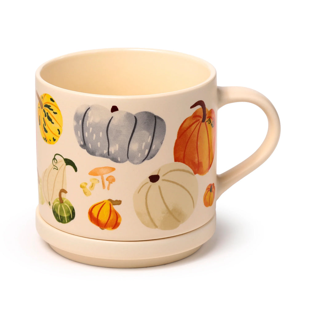 Angled view of a cream porcelain mug decorated with an autumn harvest pattern of colourful gourds, pumpkins, and mushrooms, standing on its matching coaster lid.