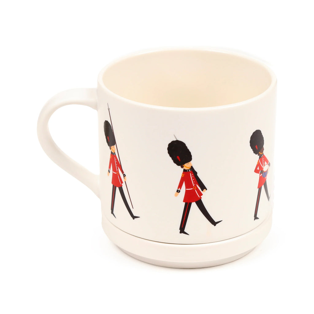 Angled view of an off-white porcelain mug, featuring a wraparound design of cartoon London Guardsmen. The guardsmen are depicted in red uniforms and black bearskin hats; one figure holds a spear, another plays a drum. The mug has a simple white handle and an off-white interior.