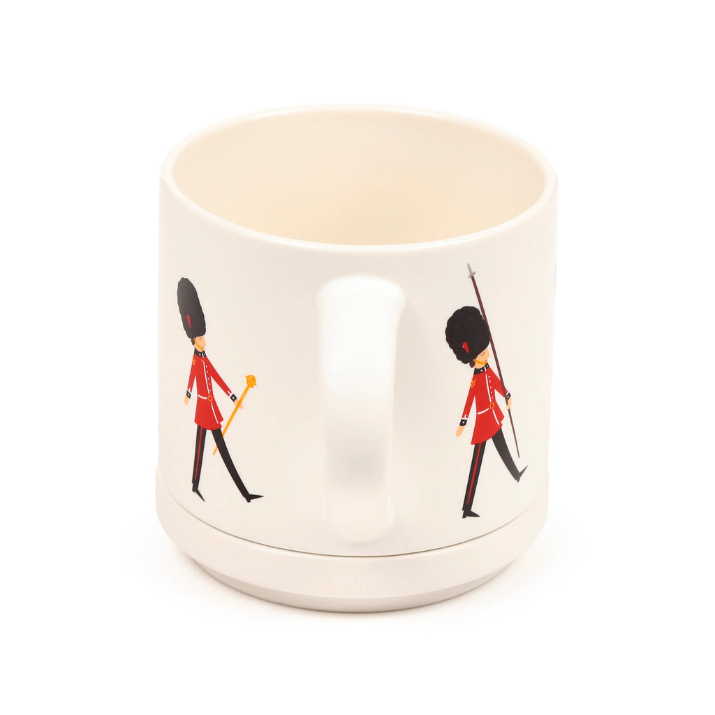 Angled view of a cream porcelain mug with a cream handle, decorated with two cartoon London Guardsmen in red uniforms and bearskin hats, resting on a matching cream coaster.