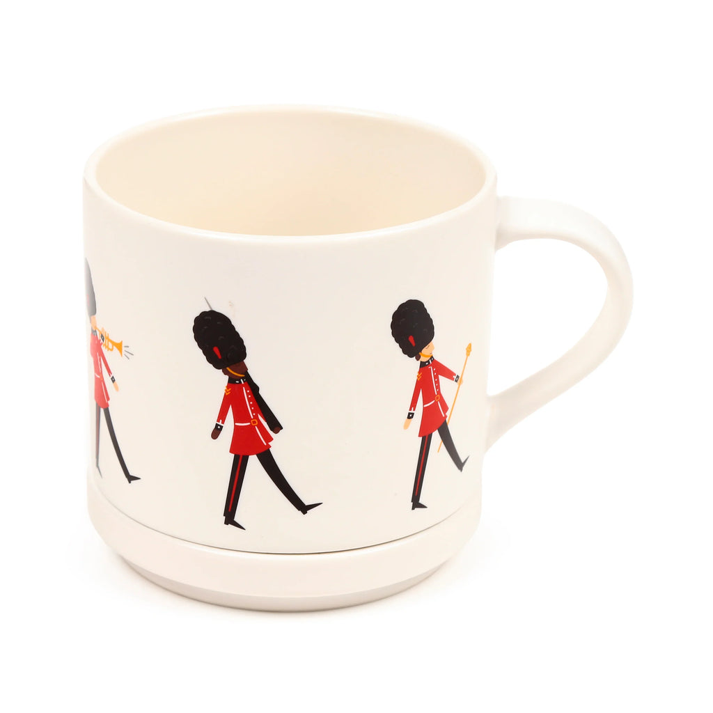 Angled view of a cream porcelain mug, featuring illustrations of London Guardsmen in red uniforms and bearskin hats, resting on its matching coaster lid, with the handle on the right.