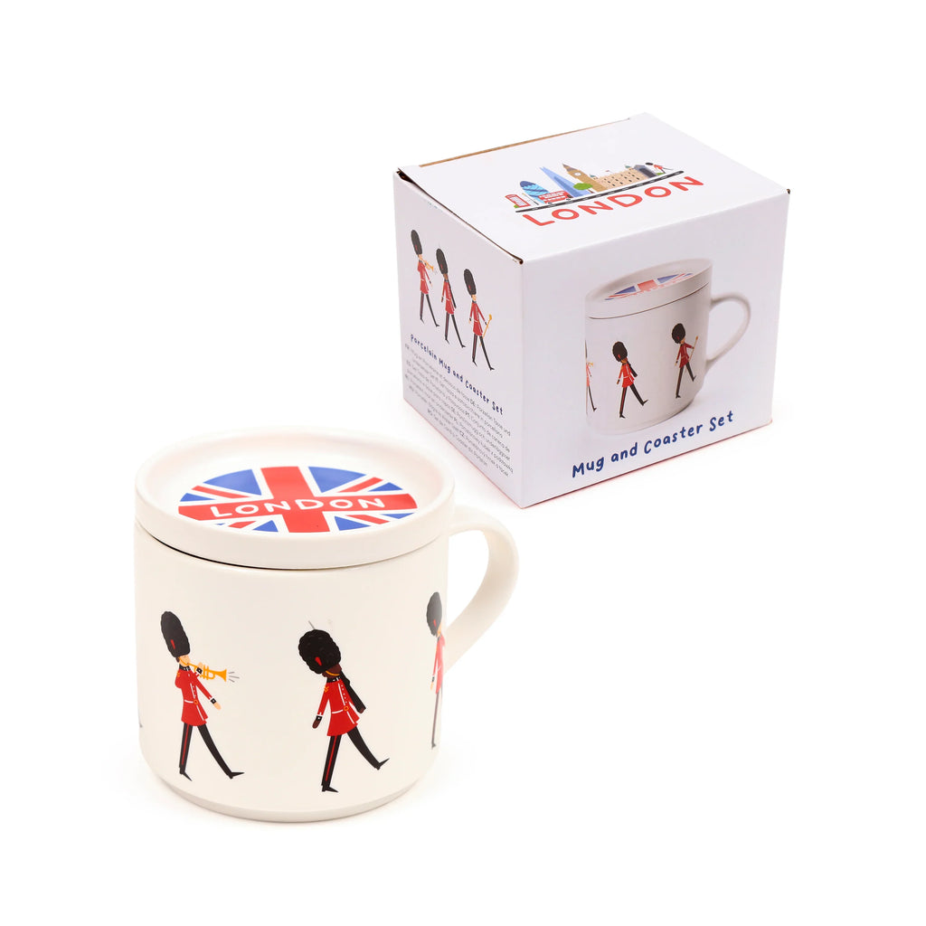 Angled view of a white porcelain mug decorated with marching London Guardsmen in red uniforms. The mug is topped with a coaster lid featuring a Union Jack flag and 'LONDON' text. A white gift box with London landmarks and Guardsmen designs is visible in the background.