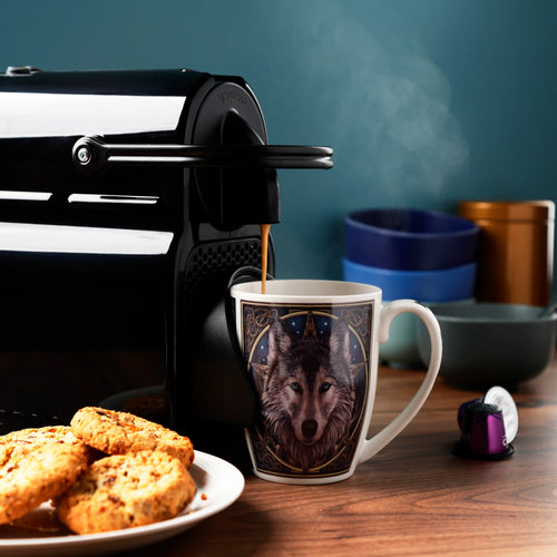 In-context lifestyle shot of a Lisa Parker Wolf Head porcelain mug, featuring a detailed illustration of a grey wolf with blue eyes surrounded by celestial patterns. The mug, currently being filled with hot coffee from a black machine, sits on a wooden counter alongside a plate of cookies and a purple coffee capsule.