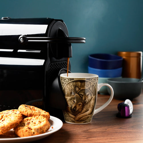 Lisa Parker Loyal Companions Wolf Porcelain Mug, featuring an intricate two-wolf and Celtic knotwork design, in an in-context lifestyle shot showing steaming coffee being poured from an espresso machine, with biscuits on a plate.