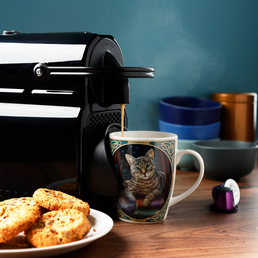 In-context lifestyle shot of a Lisa Parker Fortune Teller Cat porcelain mug, depicting a tabby cat with a crystal ball, being filled with steaming coffee from an espresso machine, next to a plate of biscuits.