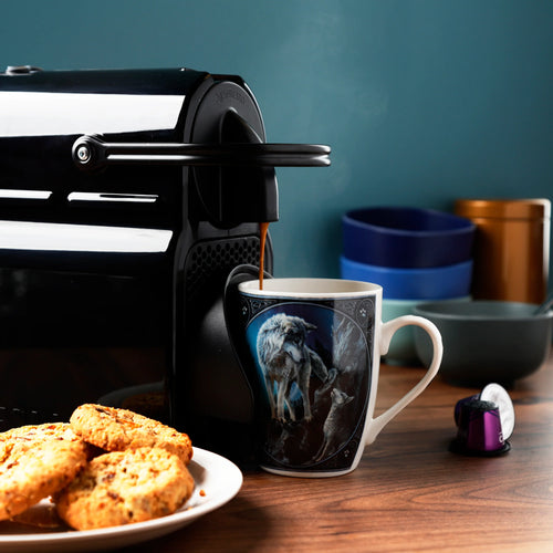 Angled, in-context shot of a Lisa Parker Guidance Wolf Design porcelain mug. The white mug features a detailed illustration of a grey wolf and a pup, with steam rising as coffee is poured into it. It sits on a wooden table next to a plate of cookies.