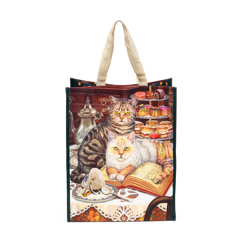 Front view of a reusable shopping bag with beige fabric handles, featuring an illustration of a tabby cat and a ginger cat at a tea party with a silver teapot, teacup, and tiered desserts. The ginger cat rests on an open book titled 'Brewed Prophecies by Lisa Parker'.