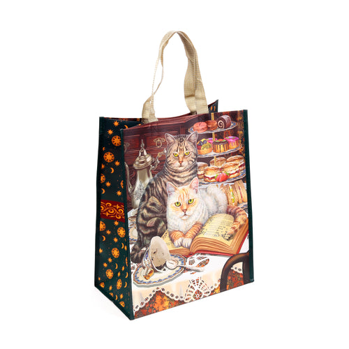 Angled view of a reusable shopping tote bag, featuring an illustration of a tabby cat and a ginger cat at a tea party with an open book titled 'Brewed Prophecies By Lisa Parker', tea set, and pastries. Dark green side panels display orange star and moon patterns.