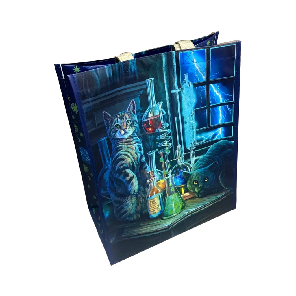 Angled view of a reusable shopping bag, depicting a tabby cat and a black cat surrounded by laboratory flasks with colourful liquids, with blue lightning visible through a window. The dark blue side panel features a pattern of green stars and moons, and beige fabric handles are visible at the top.
