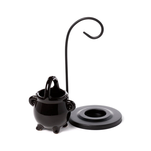 Slightly angled view of a black ceramic cauldron oil burner, featuring a top handle and three small feet, positioned beside a black metal hanging stand with a hooked arm and circular base.