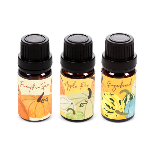 Angled view of a set of three autumn harvest fragrance oil bottles. The dark brown bottles display labels for 'Pumpkin Spice', 'Apple Pie', and 'Gingerbread' scents, adorned with colourful gourds and pumpkins.