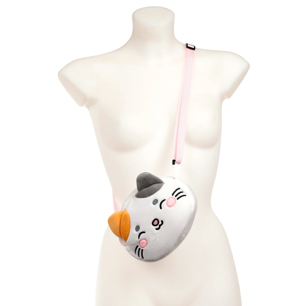 Front view of a plush white and grey cat-shaped crossbody bag with an embroidered face, contrasting dark grey and orange ears, and a pink fabric strap, displayed on a mannequin.