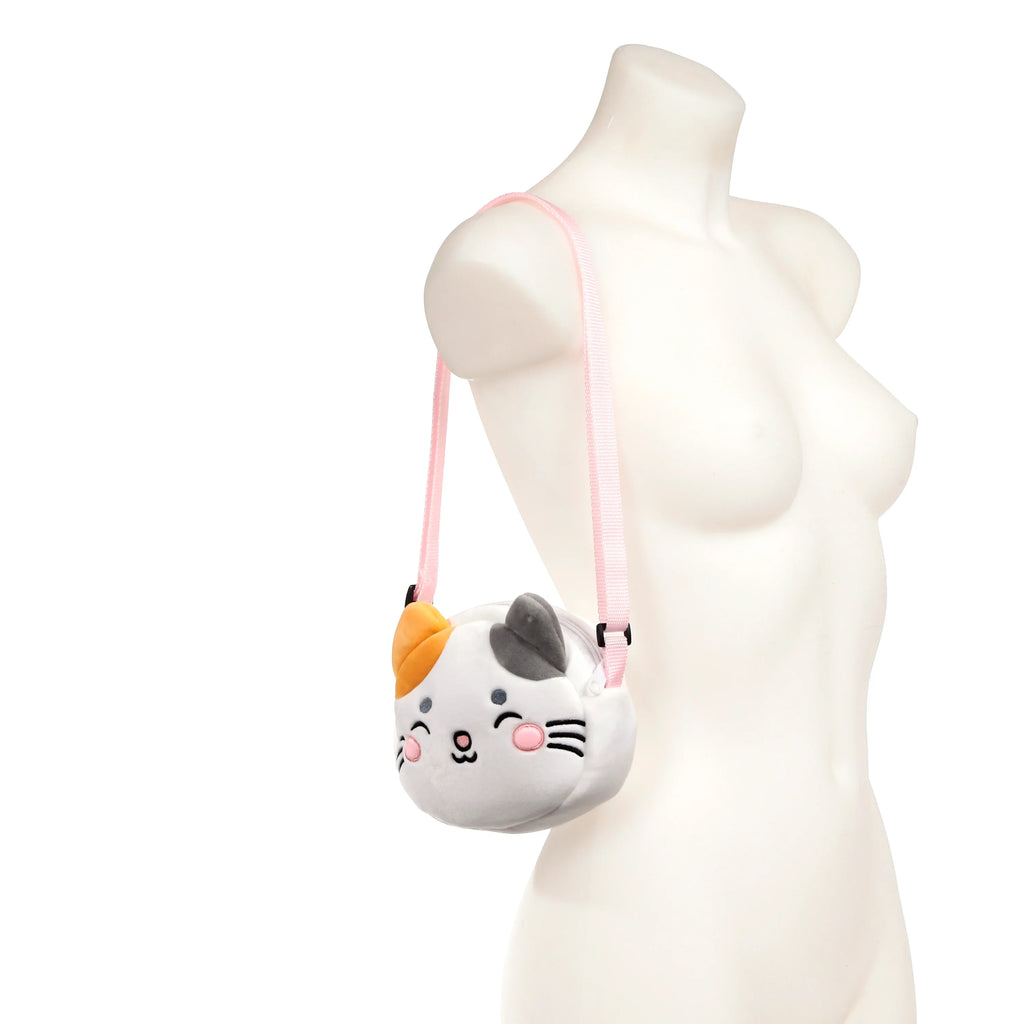 Plush crossbody bag shaped like a white, grey, and orange cat's face, with an embroidered winking eye, pink nose, and blushing cheeks, worn on a white mannequin torso with a pink adjustable strap.