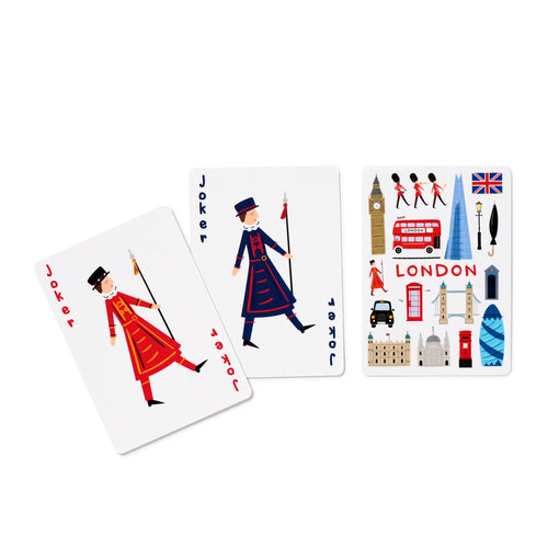 Overhead view of a London souvenir standard deck of playing cards. Two Joker cards depict Beefeaters in red and blue uniforms, while a third card features a collage of iconic London landmarks and symbols, including Big Ben, a red double-decker bus, Tower Bridge, and the Union Jack flag.