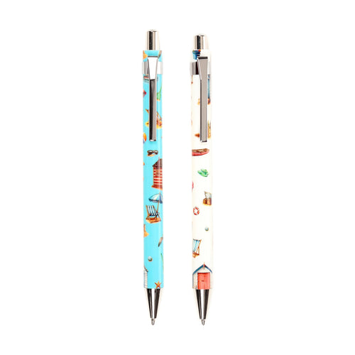 Coastal twin pen set, displaying two pens upright on a white background: one light blue with beach hut, deck chair, and sunglasses illustrations; the other cream with life buoy, flip-flop, and boat patterns.