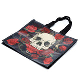Angled view of a black reusable shopping bag featuring a prominent white skull surrounded by vibrant red roses, a moth, and a swallow, with black fabric handles.