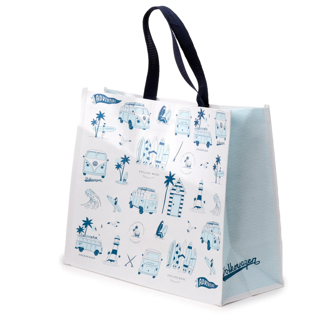 Angled view of a white reusable shopping bag with dark blue fabric handles. The front features a blue beach-themed pattern, including Volkswagen T1 camper vans, surfboards, palm trees, a lighthouse, and text like 'Explore More'. The visible side of the bag is light blue with a subtle wavy pattern.