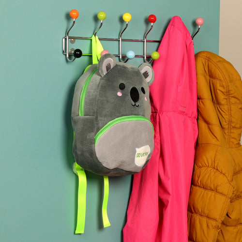 Front view of a grey plush koala rucksack backpack with bright green zips and straps, featuring embroidered koala face details and a 'SQUEAK' patch. The backpack is shown hanging on a multi-coloured coat rack in a lifestyle setting, alongside a bright pink jacket and a mustard-coloured coat.