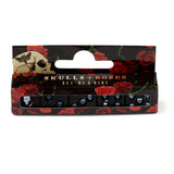 Front view of a set of five black dice, each with a white skull design, presented in a display box decorated with red roses and a skull and moth illustration.