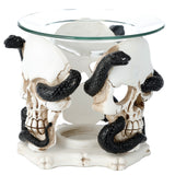 Angled view of a white resin skull and serpent oil burner, featuring two aged skulls with black coiled snakes and a clear glass dish on top.