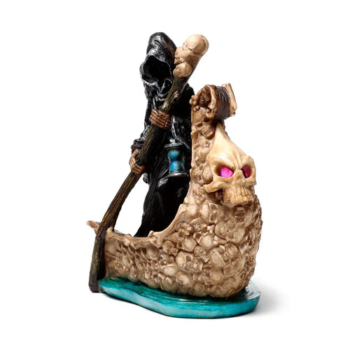 Angled view of a decorative LED 'Reaper Boat of Skulls' ornament, featuring a black-robed Grim Reaper figurine holding a skull-topped staff and hourglass. The boat is intricately detailed with numerous beige skulls, topped by a large skull with vibrant pink eyes at the front, all resting on a turquoise base.