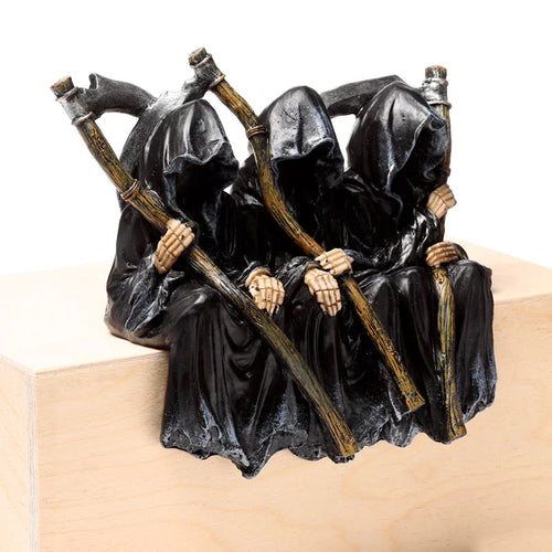 Angled view of a dark decorative ornament featuring three hooded Grim Reaper figures in black robes, with skeletal hands holding scythes, depicted seated.