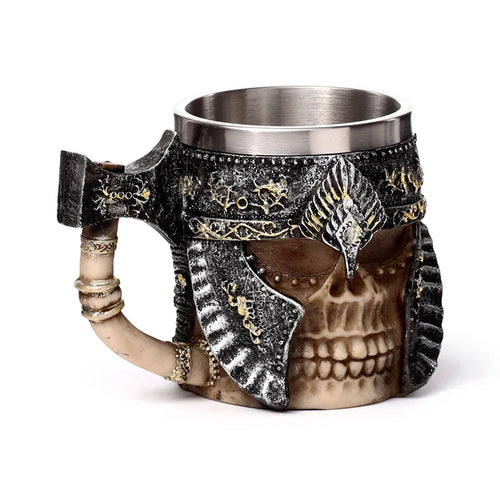Angled view of a decorative tankard featuring an armoured skull design, with a dark grey and gold helmet, bone-coloured skull, and an axe-shaped handle. The tankard has a silver-coloured metallic interior.