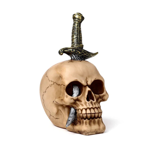 Angled view of a decorative, bone-coloured skull ornament with a gold-hilted sword piercing the top and emerging from the eye socket.