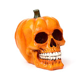 Angled front view of an orange decorative pumpkin skull ornament, featuring hollow eye sockets, a nose cavity, and prominent white teeth, with a green stem and ridged pumpkin texture.