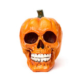 Front view of an orange pumpkin skull decorative ornament, featuring hollow eye sockets, a prominent set of white teeth, and a green stem on top.