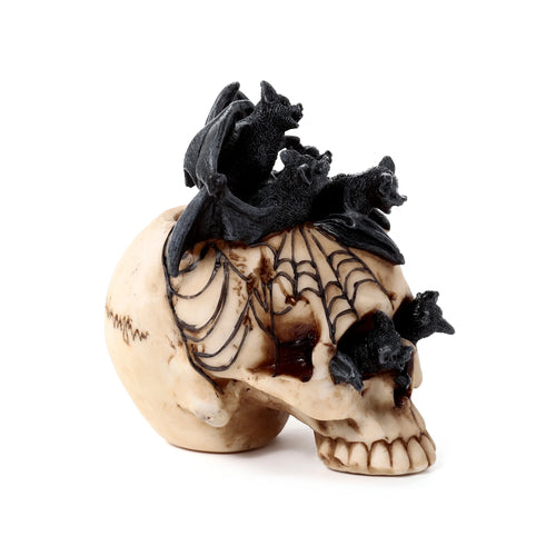 Angled view of a light-coloured decorative skull ornament, depicting black bats crawling on top and emerging from the eye socket, with sculpted spiderweb details and visible skeletal sutures.