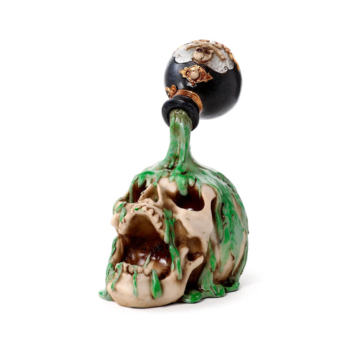 Front view of a decorative screaming skull ornament, beige in colour, with green potion dripping down from an ornate black bottle atop its head. The bottle features intricate gold and silver-toned detailing.