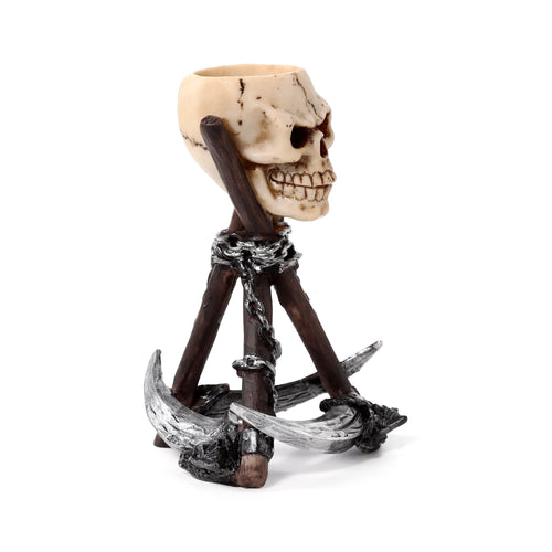 Angled view of a resin tea light holder, featuring a beige skull head perched on a dark, chained tripod base with silver-coloured scythe blades forming the feet.