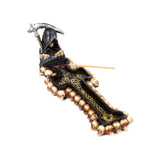 Angled view of a decorative incense burner, depicting a hooded Grim Reaper figure holding a scythe at one end of a black cross adorned with numerous small skulls and gold decorative patterns, with a pale wood incense stick with a purple tip inserted.