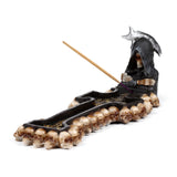 Angled view of a decorative cross-shaped Reaper incense burner, featuring a hooded Grim Reaper figure holding a scythe, with skeletal hands and rows of skulls along its edge, with an unlit incense stick.