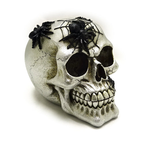 Silver skull ornament, angled to show its top, right side, and front, adorned with black spiders and webs, featuring visible eye sockets, nasal cavity, and prominent teeth.