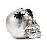 Decorative silver skull ornament with black spiders and web patterns, shown from an angled right profile, revealing its textured finish and teeth.