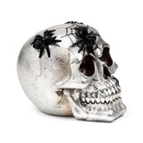 Angled view of a decorative silver skull ornament, featuring black spiders and spider webs on its textured metallic surface. The visible teeth and dark eye sockets are prominent.