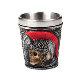 Decorative shot glass, angled view, featuring a Roman soldier helmet design with a prominent red plume, a skull face, and distressed black, silver, and gold detailing, with a metallic inner lining.