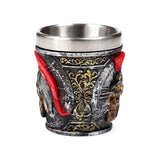 Angled view of a decorative shot glass with a metallic silver inner cup, featuring an outer design of a weathered dark grey and black Roman soldier's helmet with a visible skull, red plume, and ornate gold patterns.