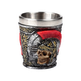 Decorative shot glass, showing a textured dark grey and black body with gold detailing, featuring a skull wearing a Roman soldier helmet with a prominent red crest, and a silver-coloured metal interior.