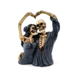 Front view of a decorative skeletal couple ornament, with their bone-coloured arms raised to form a heart shape. The female skeleton has dark hair and a purple flower, and the male skeleton wears a dark open robe.