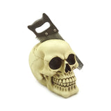 Angled view of a bone-coloured decorative skull ornament, featuring a dark-handled saw passing through its head. The ornament shows detailed eye sockets, nasal cavity, and teeth, all on a plain white background.