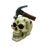 Angled view of an off-white decorative skull ornament with a dark hammer resting on its cranium and several silver nails embedded, presented on a white background.