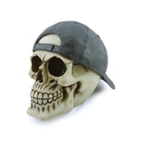 Angled view of a decorative skull ornament, featuring a light beige skull with dark eye sockets and detailed teeth. It wears a dark grey baseball cap backwards, with its adjustment strap visible at the rear.