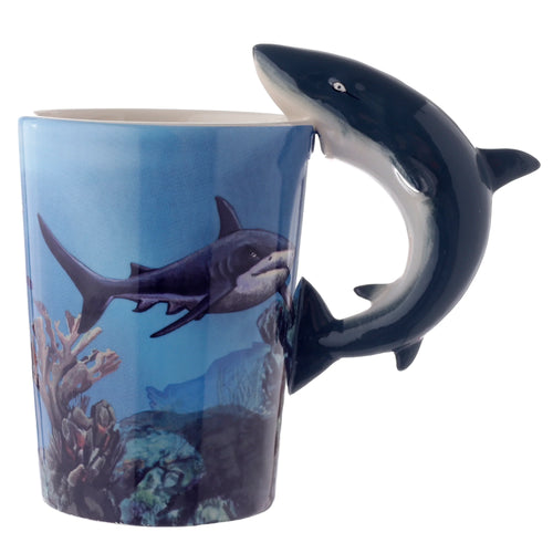 Angled view of a ceramic mug with a dark blue/grey shark-shaped handle. The mug features a blue underwater scene depicting a shark and coral reef, with a white interior.