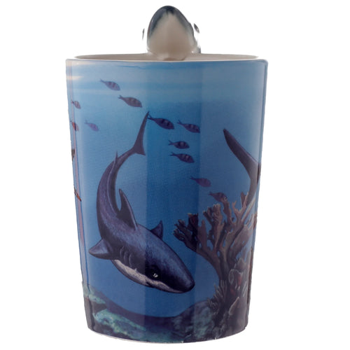 Angled view of a blue ceramic mug depicting an underwater scene with a large shark, smaller fish, and coral, featuring a shaped handle formed by a shark's head.