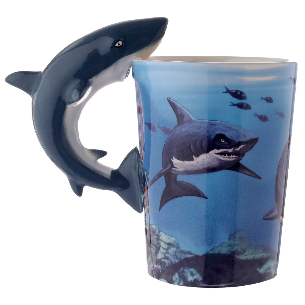 Angled view of a ceramic mug with a dark blue shark-shaped handle. The mug features an illustration of a shark swimming in blue ocean water, accompanied by smaller fish and rocky seabed details.