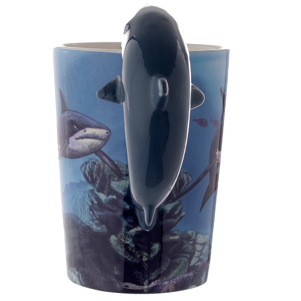 Ceramic mug from an angle highlighting its glossy, dark grey shark-shaped handle. The mug's blue surface features an underwater design with sharks and coral.